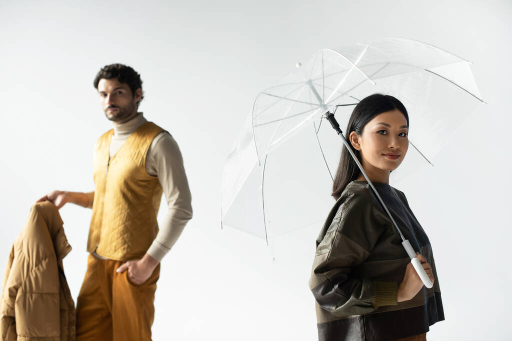 femme asiatique souriant à la caméra sous parapluie transparent près de l'homme tenant veste gonflante sur gris - Photo, image