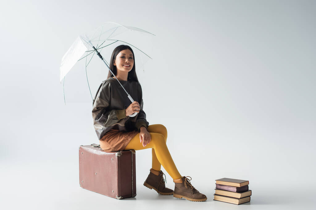 счастливая азиатка с прозрачным зонтиком сидит на винтажном чемодане рядом с книгами на сером - Фото, изображение