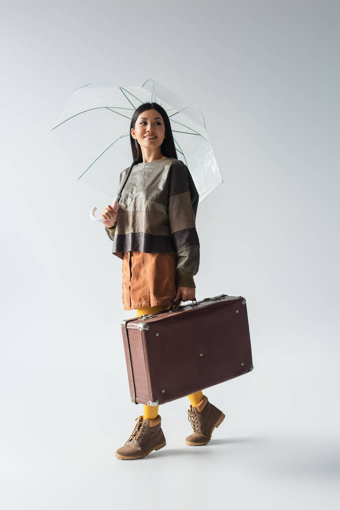 volle Länge Ansicht der stilvollen asiatischen Frau mit Vintage-Koffer zu Fuß unter transparenten Regenschirm auf grau - Foto, Bild