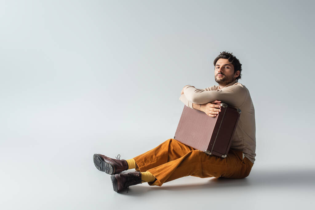 Ganzkörperansicht eines Mannes in trendiger Kleidung, der mit einem Vintage-Koffer auf grau sitzt - Foto, Bild