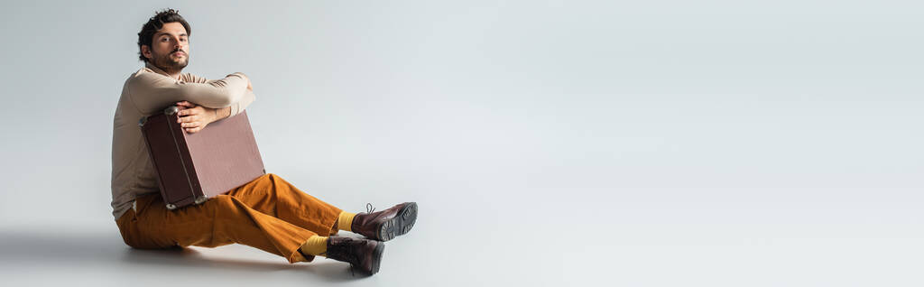 visão comprimento total do homem em calças amarelas e botas marrons sentado com mala retro em cinza, banner - Foto, Imagem