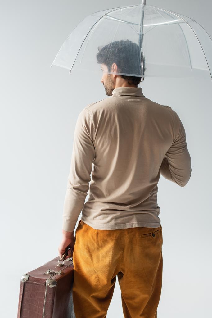 back view of stylish man with vintage suitcase standing under transparent umbrella isolated on grey - Photo, Image
