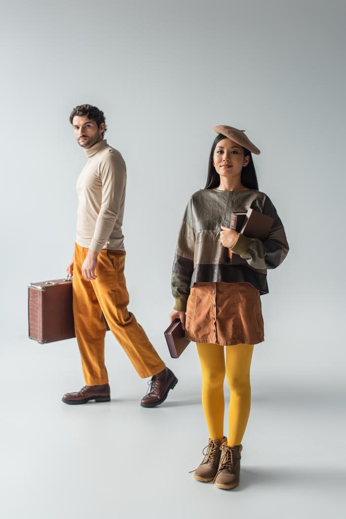 full length view of man with vintage suitcase and asian woman with books on grey - Foto, Imagem