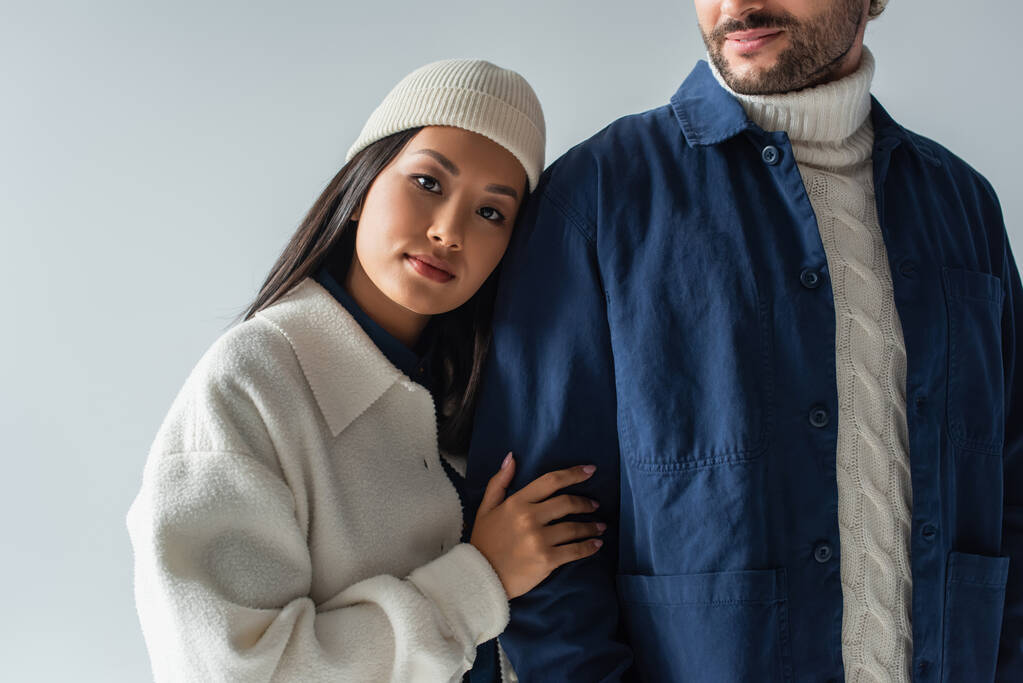 giovane donna asiatica in berretto appoggiata sull'uomo in giacca blu isolata sul grigio - Foto, immagini