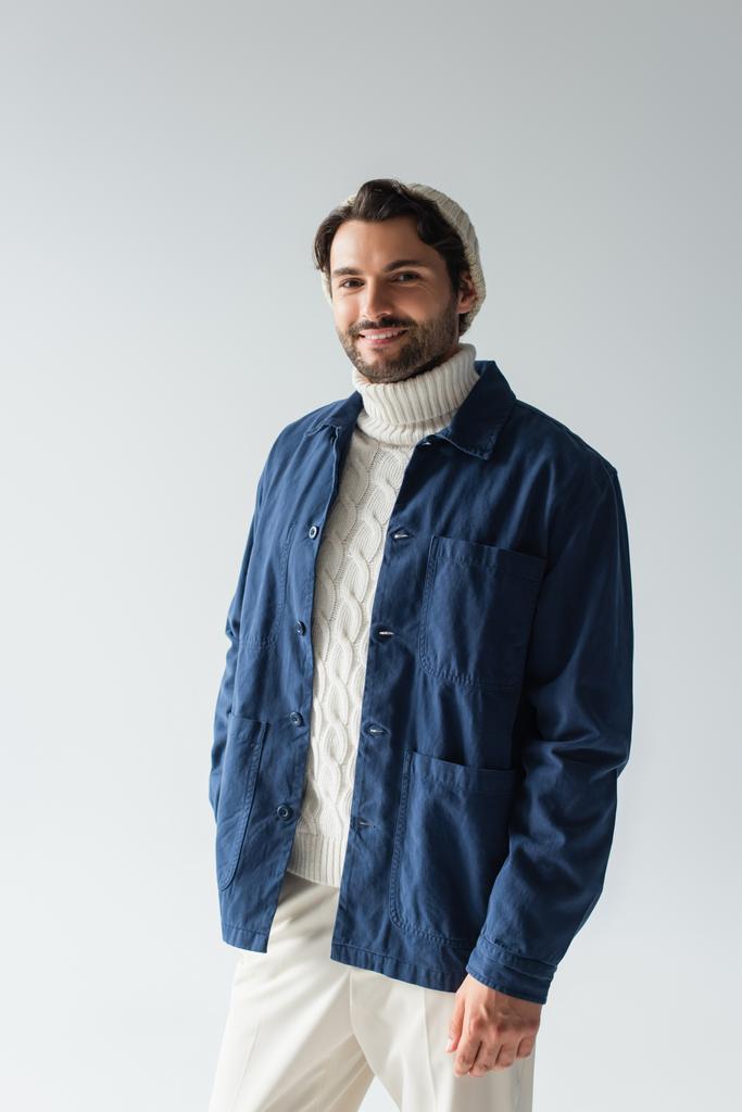 счастливый мужчина в синей куртке и белом трикотажном свитере, улыбающийся в камеру, изолированную на сером - Фото, изображение