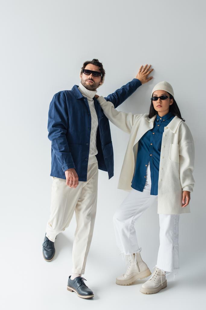 Ansicht des stilvollen gemischtrassigen Paares in weiß-blauer Kleidung und Sonnenbrille auf grau - Foto, Bild