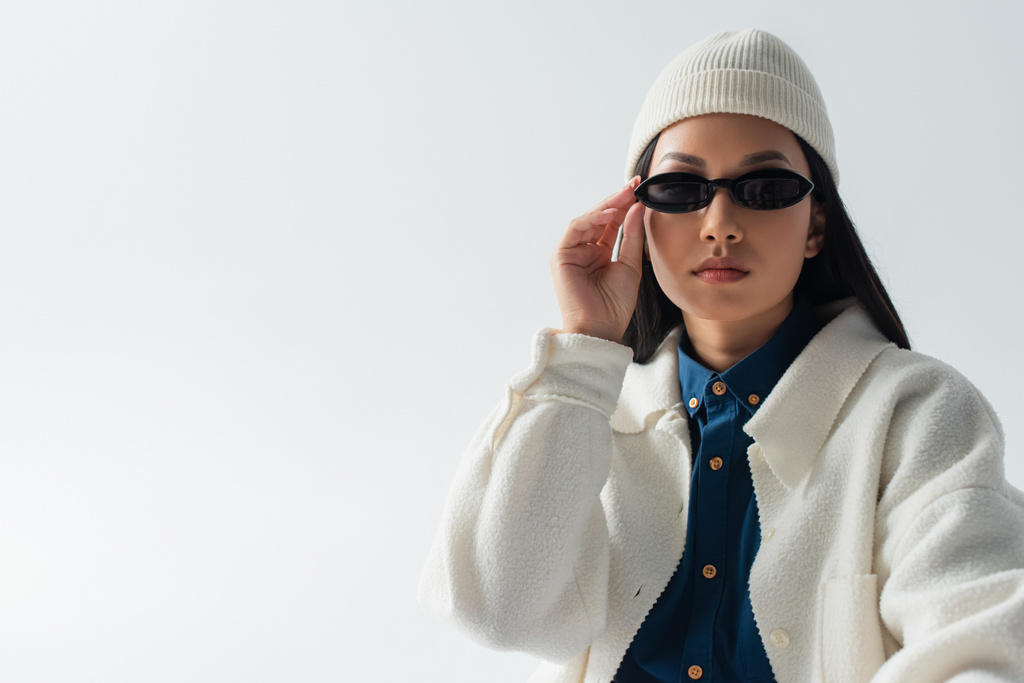 fiatal ázsiai nő fehér kabát és sapka beállítása sötét napszemüveg elszigetelt szürke - Fotó, kép