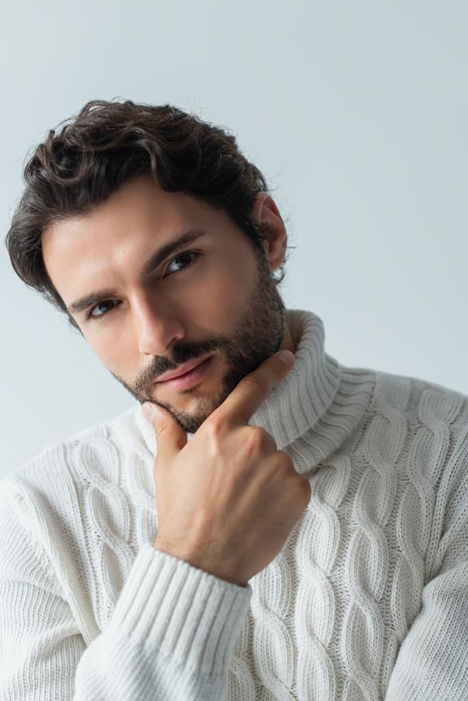brunette man in white knitted sweater touching chin and looking at camera isolated on grey - Photo, Image