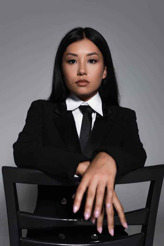 joven asiático mujer en negro formal desgaste mirando cámara mientras posando en silla aislado en gris - Foto, Imagen