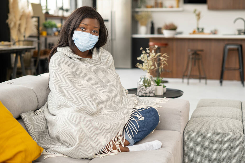 Celkový pohled na mladou nemocnou ženu v ochranné masce obličeje, která se doma necítí dobře, jak sedí na gauči přikrytá pokrývkou a cítí zimnici a horečku. Žena má nemoc - Fotografie, Obrázek