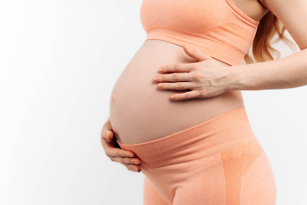 Крупним планом вагітна жінка торкається її живота на білому тлі. Жінка очікує на дитину, щаслива вагітна жінка з великим животом. Поняття вагітності, материнства, підготовки та очікування
 - Фото, зображення
