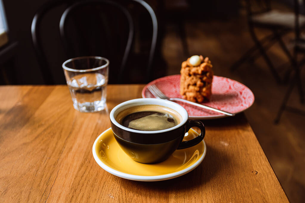 A ceramic cup of black coffee,a dessert served on a plate and a glass of water on the wooden table in a cafe. - Photo, Image