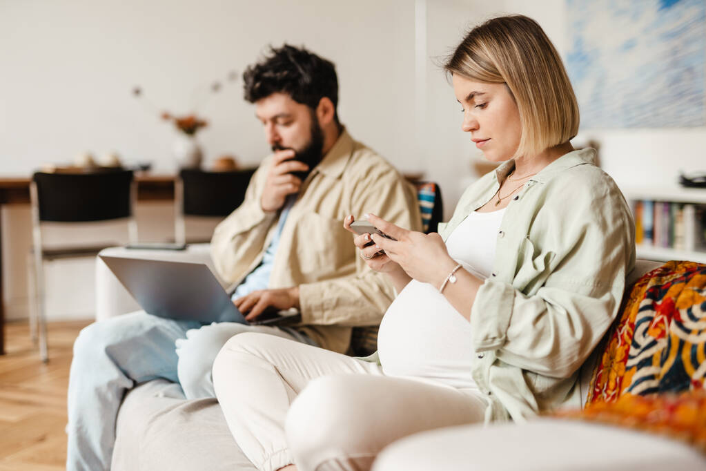 Szakállas férfi és terhes nő mobiltelefonnal és laptoppal, miközben otthon ül a kanapén - Fotó, kép