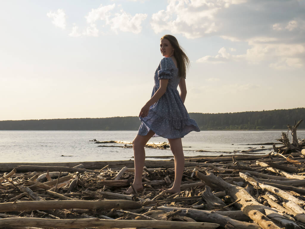 Молодая красивая босиком женщина с стройными ногами в летнем голубом платье-сарафане на берегу морской бухты на закате, концепция пляжного отдыха, летние прогулки на природе, активный образ жизни, выходные на море - Фото, изображение