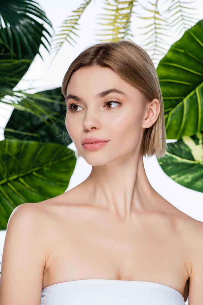 молодая женщина с идеальной кожей, отводящая взгляд от зеленых пальмовых листьев на белом - Фото, изображение