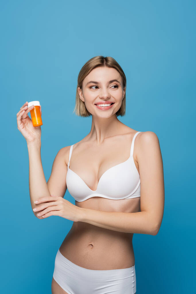 joyeuse jeune femme en lingerie blanche tenant bouteille avec des vitamines isolées sur bleu  - Photo, image