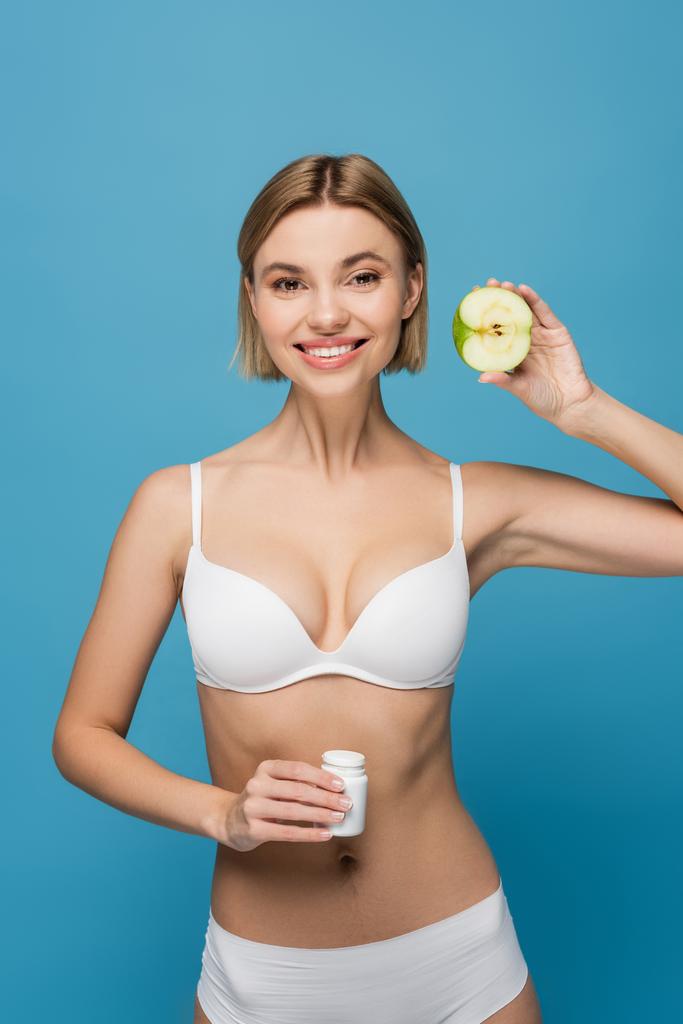 веселая женщина в белом белье держит бутылку с витаминами и яблоком наполовину изолированы на синий  - Фото, изображение