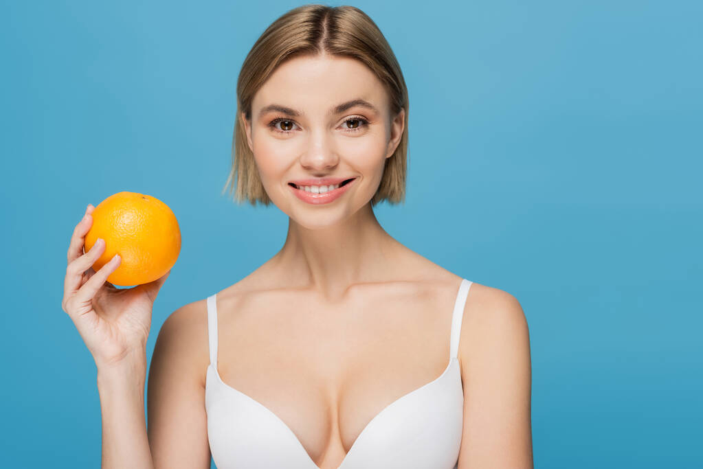 joyful young woman in white bra holding ripe orange isolated on blue  - Photo, Image