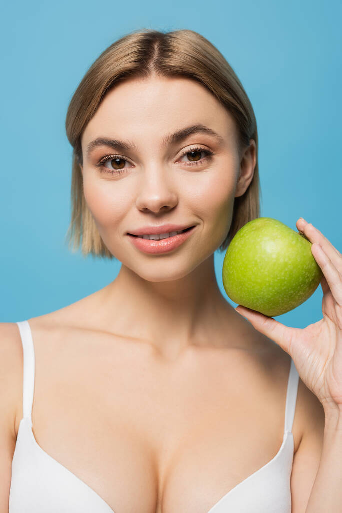 blonde young woman in bra holding green apple and smiling isolated on blue  - Photo, Image
