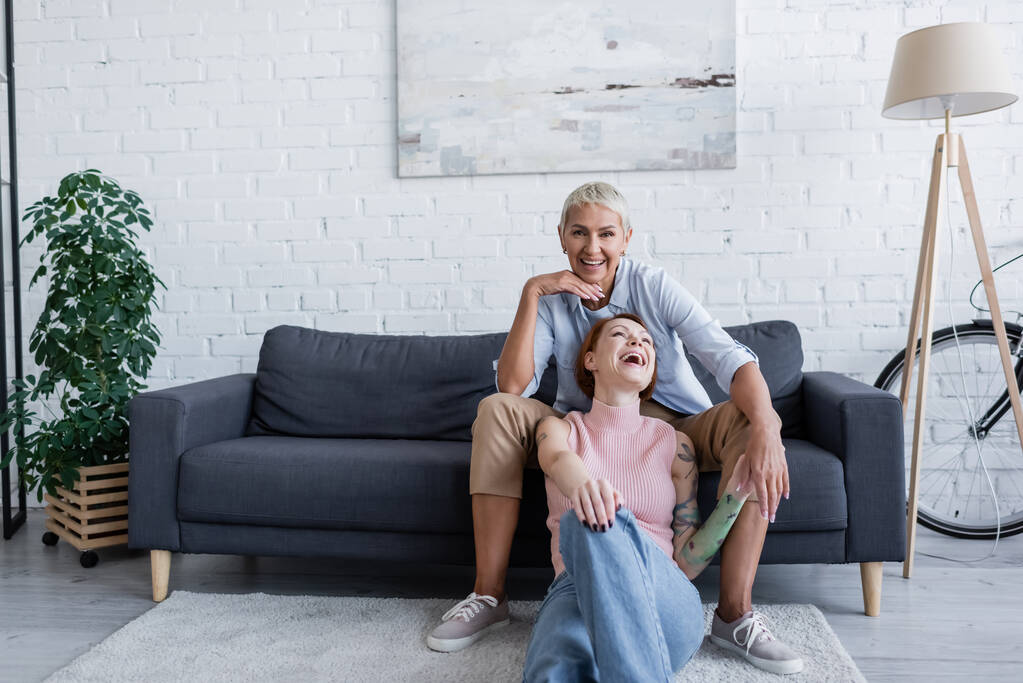 happy lesbian woman smiling at camera near girlfriend laughing on floor in living room - Photo, Image