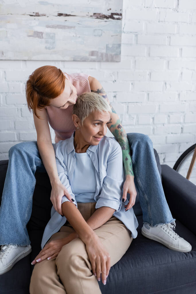tattooed woman hugging lesbian girlfriend looking away on couch at home - Photo, Image