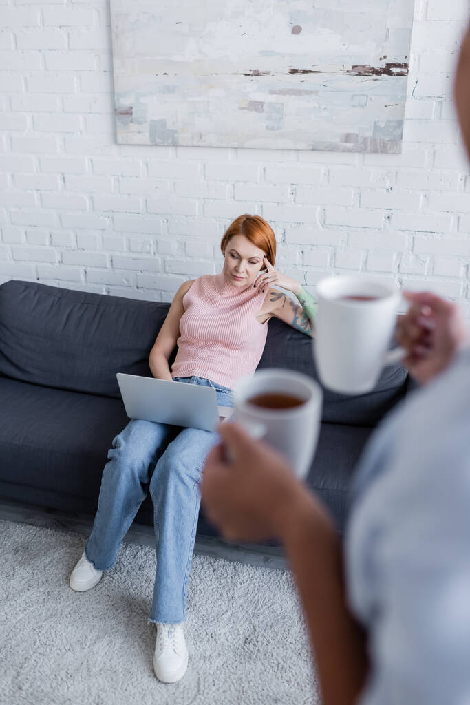 blurred woman holding cups with tea near thoughtful girlfriend sitting on couch with laptop - Photo, Image