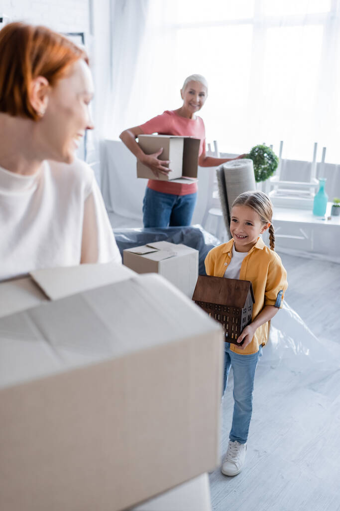 joyful dívka nesoucí dům model v blízkosti rozmazané stejné pohlaví rodiče s kartónové krabice - Fotografie, Obrázek