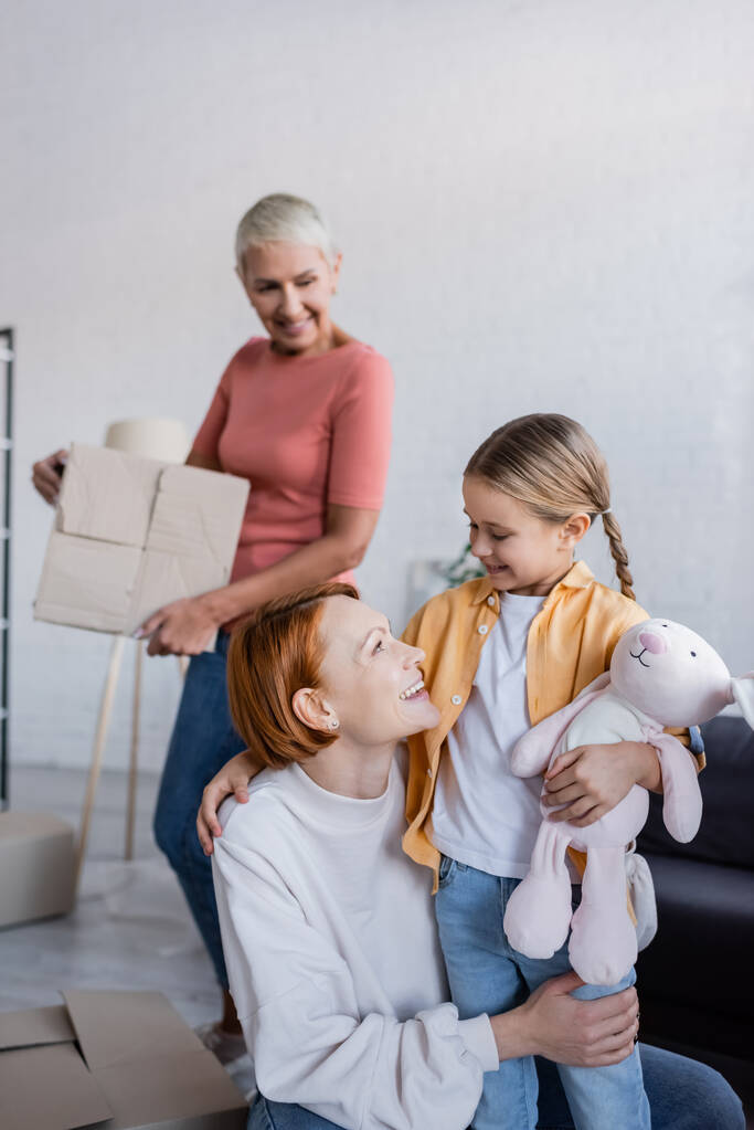 happy lesbian woman looking at adopted girl holding toy bunny in new apartment - Photo, Image