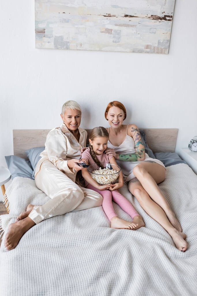 Positif lesbiennes femmes regarder la télévision près de l'enfant avec du pop-corn à la maison  - Photo, image