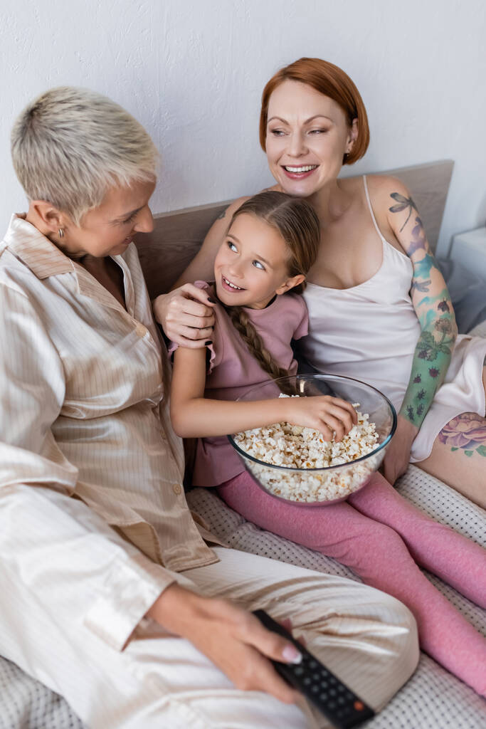 Lesbian couple hugging kid and holding remote controller on bed  - Photo, Image