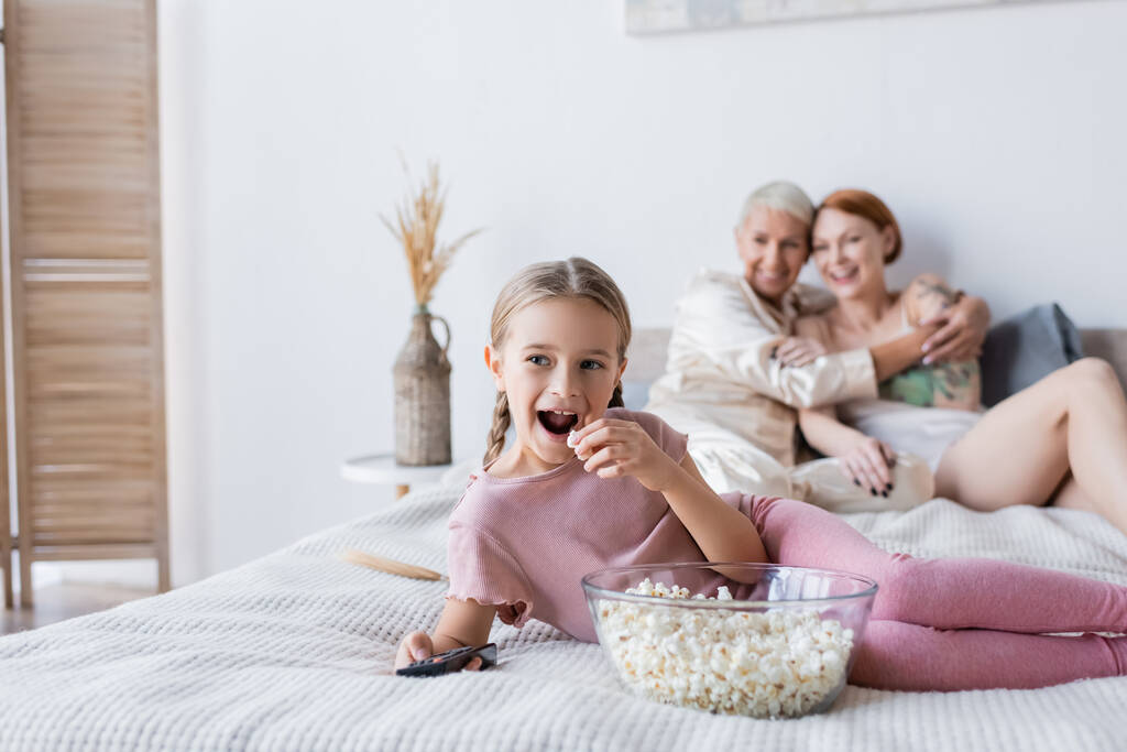 Ragazzo che tiene popcorn mentre guarda film vicino alle madri sul letto  - Foto, immagini