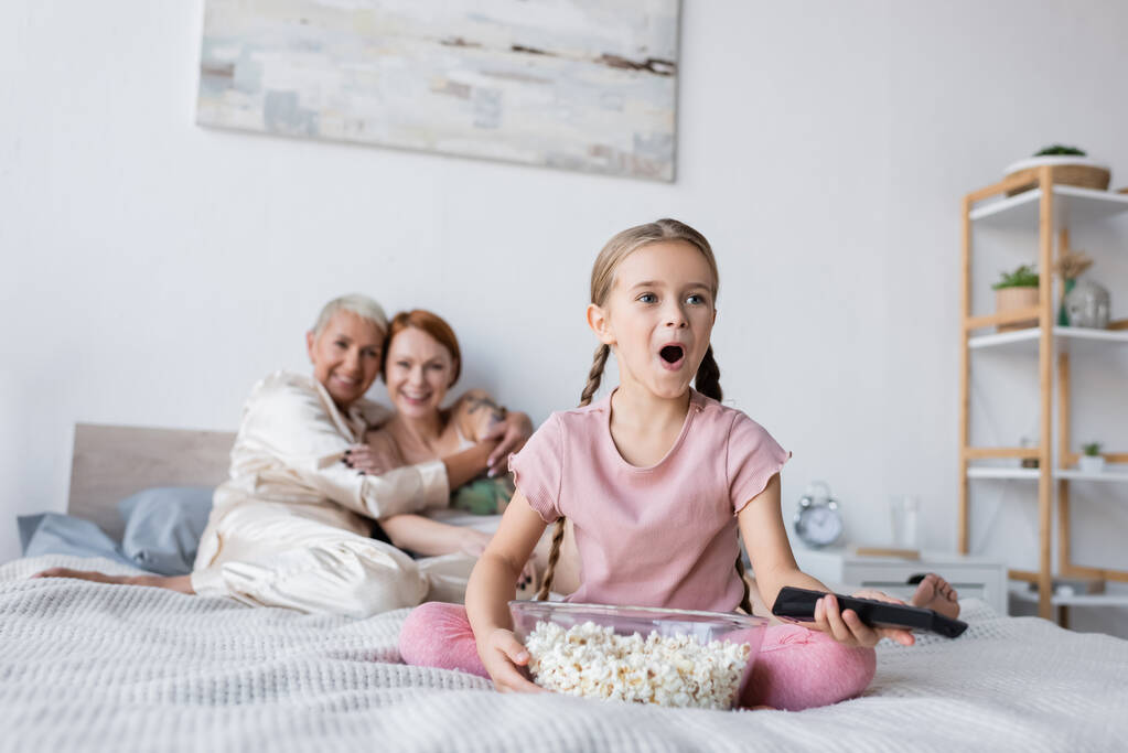 Ragazza eccitata con telecomando e popcorn vicino alle madri offuscate a casa  - Foto, immagini