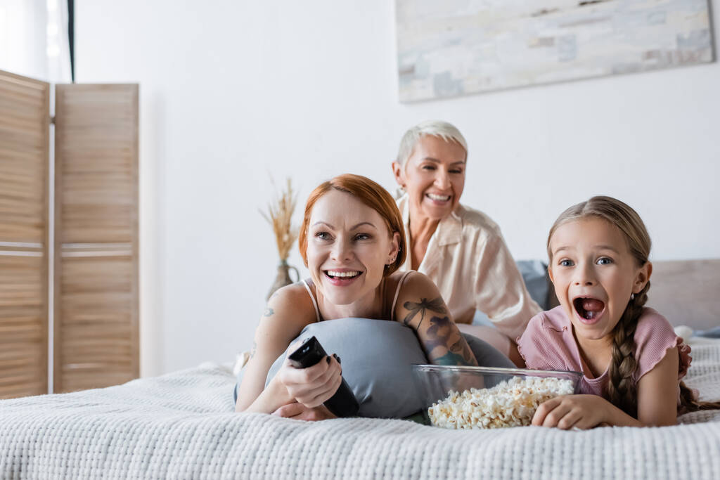Ενθουσιασμένο παιδί που βλέπει ταινία κοντά σε ποπ κορν και μητέρες στο κρεβάτι  - Φωτογραφία, εικόνα