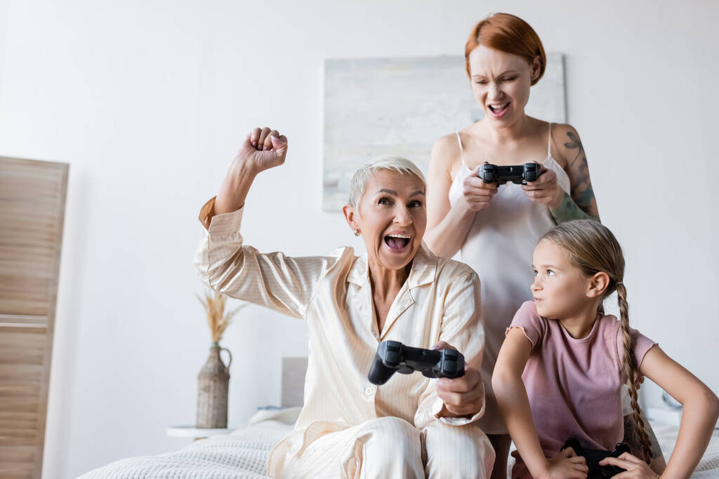 KYIV, UKRAINE - DECEMBER 8, 2021: Excited lesbian woman holding joystick near girlfriend and kid at home  - Photo, Image