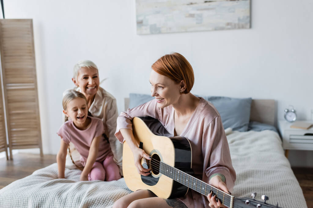 Mulher feliz tocando guitarra acústica perto de criança borrada e namorada na cama  - Foto, Imagem