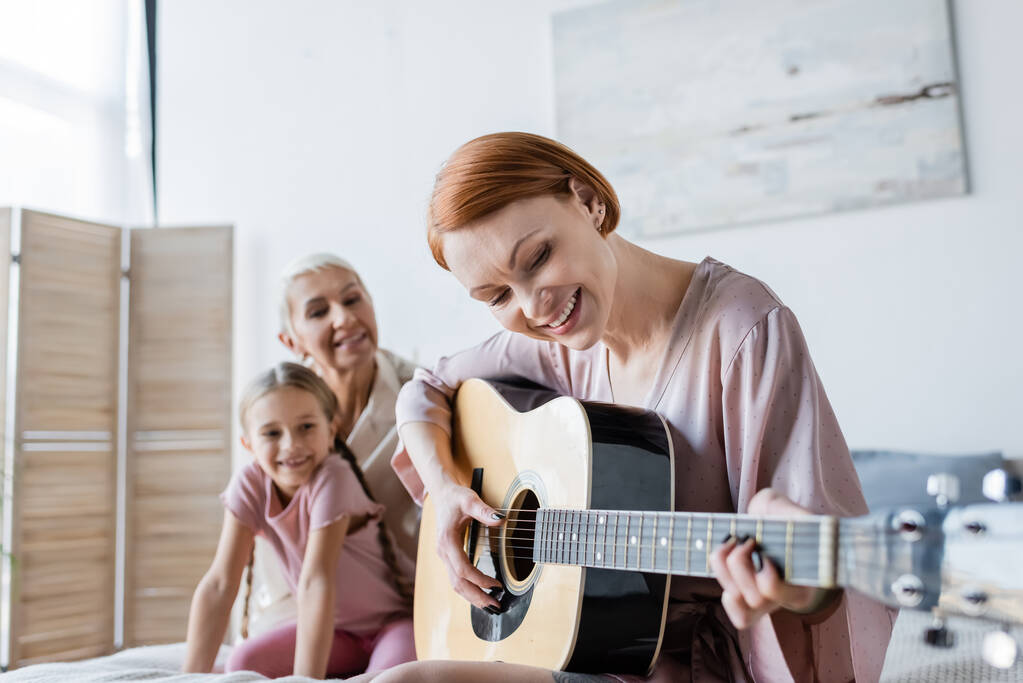 Позитивная лесбиянка, играющая на акустической гитаре рядом с размытой девушкой и ребенком дома  - Фото, изображение