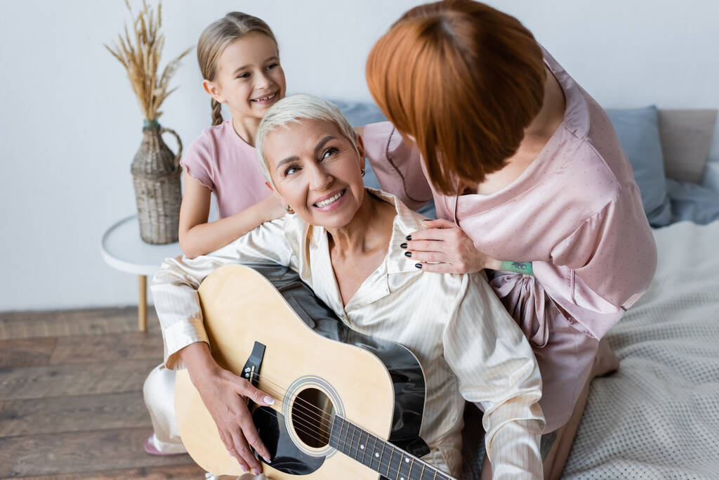 Χαμογελαστή γυναίκα παίζει ακουστική κιθάρα κοντά υιοθετημένη κόρη και φίλη στην κρεβατοκάμαρα  - Φωτογραφία, εικόνα