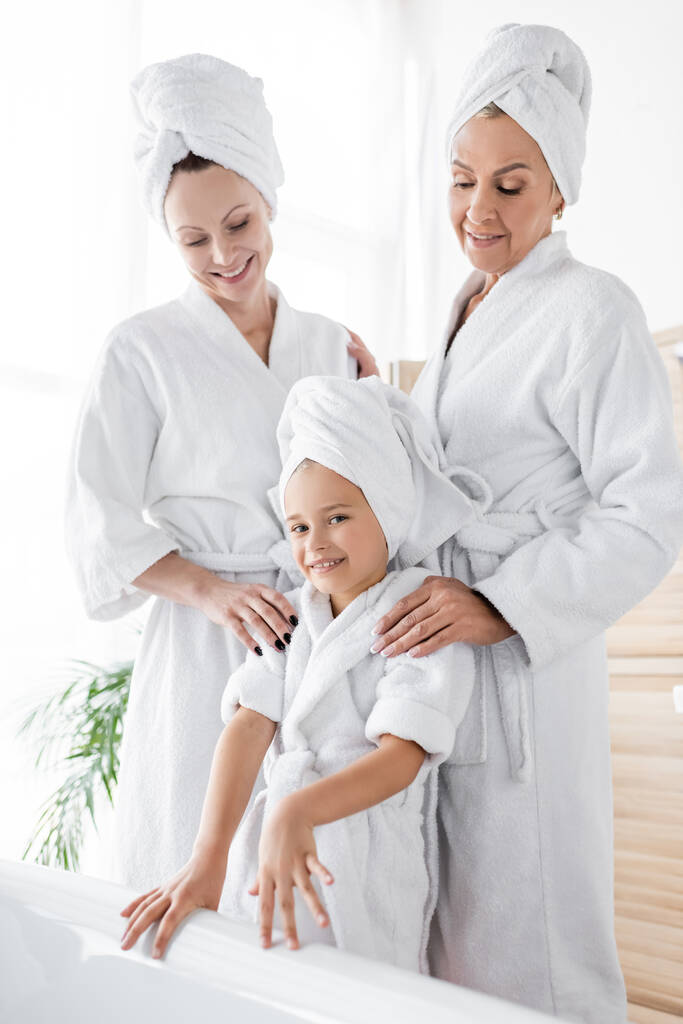Lesbian women in bathrobes standing near daughter and bathtub at home  - Photo, Image