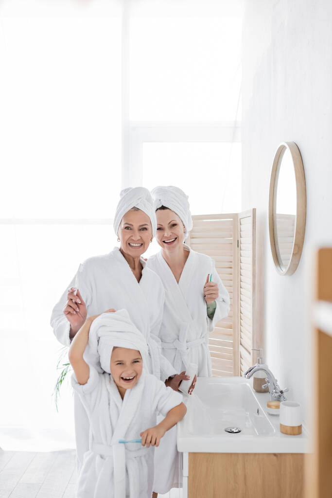 Alegres padres lesbianas en albornoces sosteniendo cepillos de dientes cerca de la hija en el baño  - Foto, Imagen