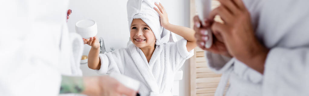 Χαμογελαστό κορίτσι με μπουρνούζι που κρατάει κρέμα κοντά σε θολές μαμάδες στο μπάνιο, πανό  - Φωτογραφία, εικόνα