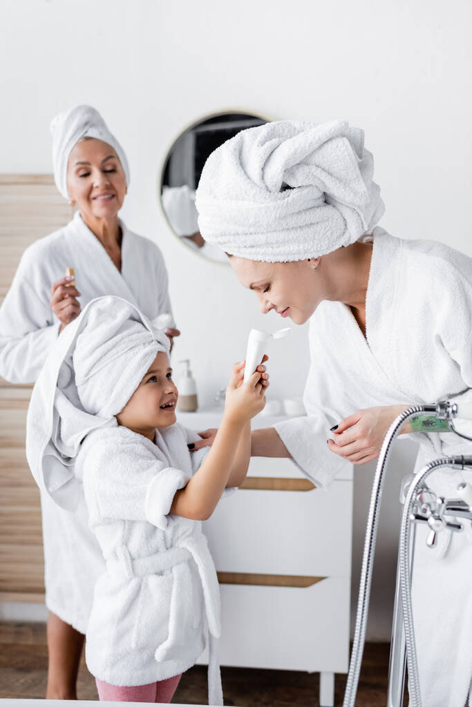 Enfant souriant en peignoir tenant la crème près des parents lesbiennes dans la salle de bain  - Photo, image