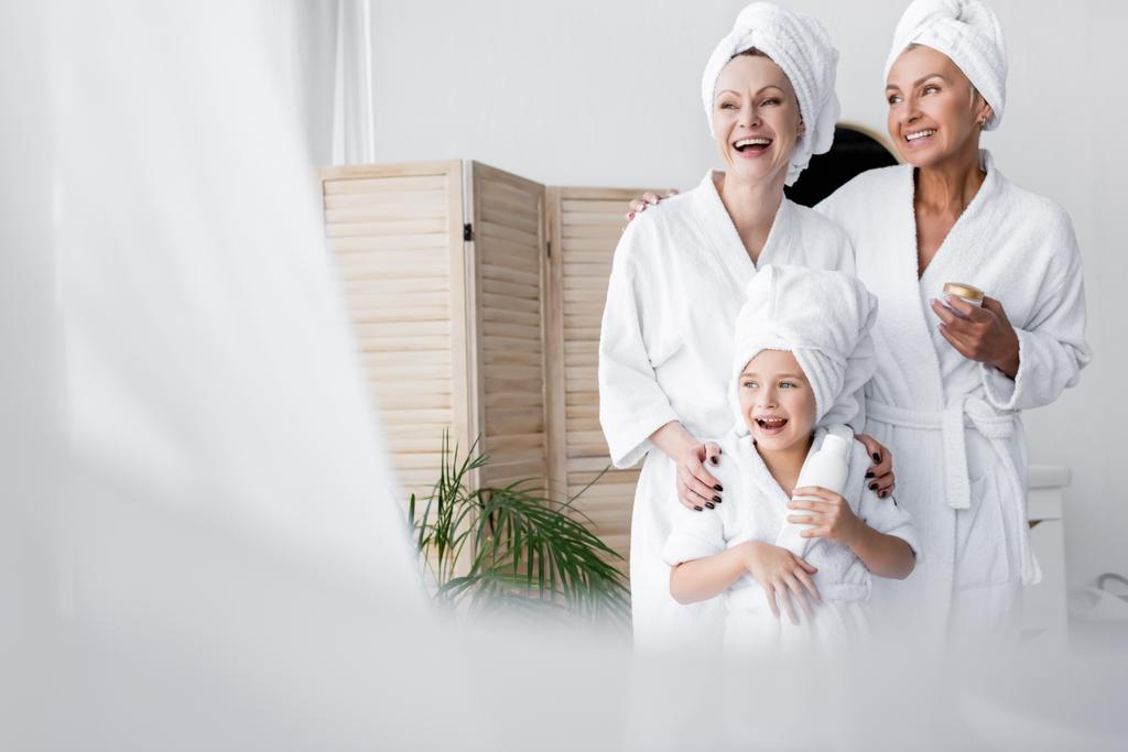 Cheerful lesbian couple and adopted daughter in bathrobes holding cosmetic cream in bathroom  - Photo, Image