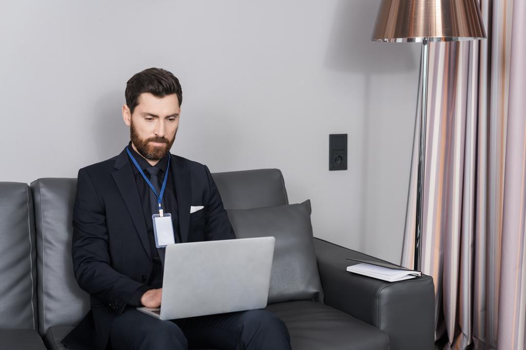 homem de negócios barbudo com crachá de identificação usando laptop enquanto sentado no sofá de couro no quarto do hotel - Foto, Imagem