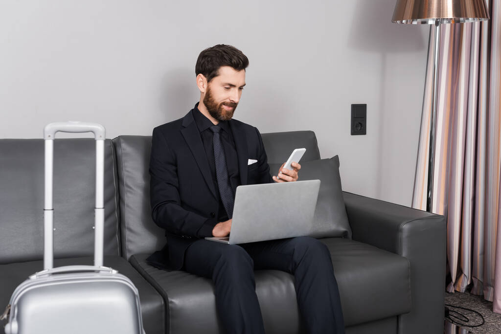 fröhlicher Geschäftsmann mit Smartphone in der Nähe von Laptop und Gepäck im Hotelzimmer - Foto, Bild
