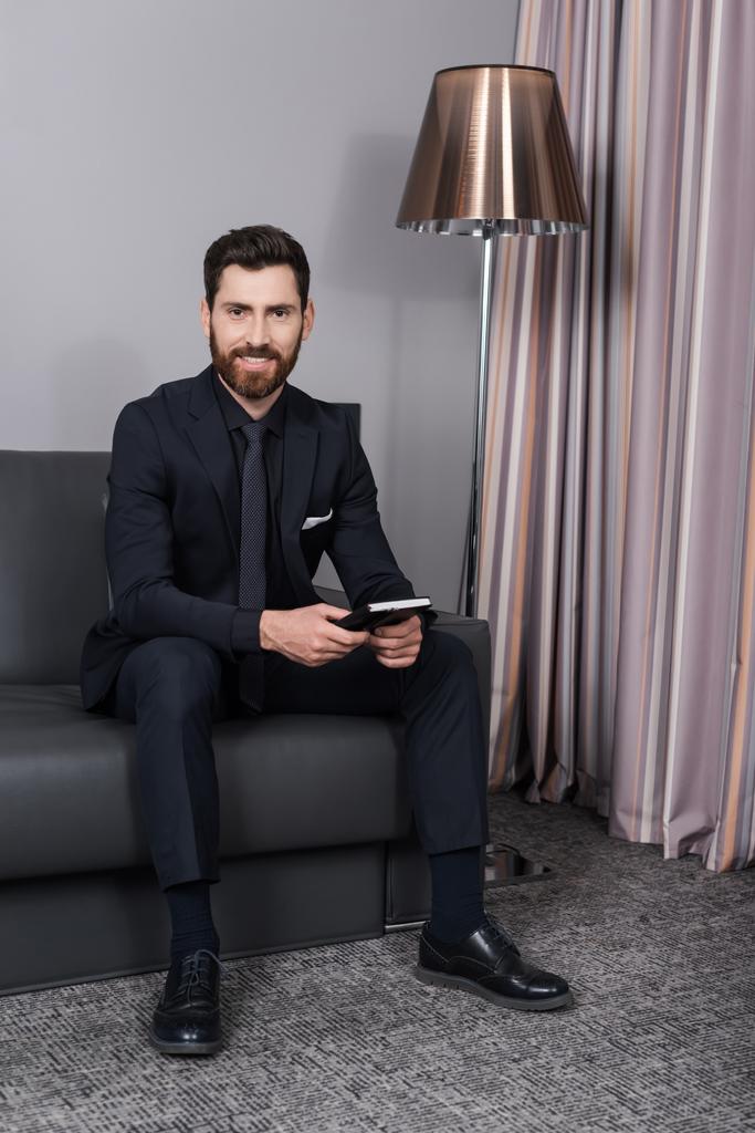 happy man in suit holding notebook and sitting on leather couch in hotel room - Photo, Image