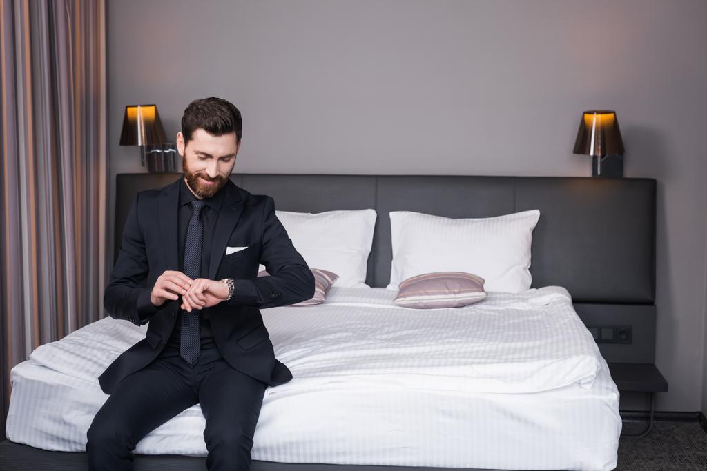 hombre alegre en traje comprobar el tiempo mientras está sentado en la cama en la habitación de hotel  - Foto, imagen