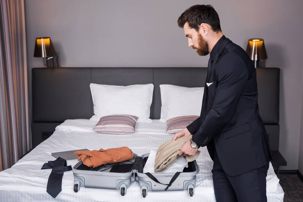 zijaanzicht van de man met de baard in pak die zijn koffer uitpakt in de hotelkamer  - Foto, afbeelding
