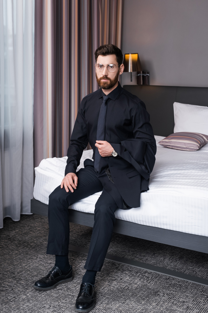bärtiger Geschäftsmann mit Brille und Anzug sitzt auf Bett im Hotelzimmer  - Foto, Bild