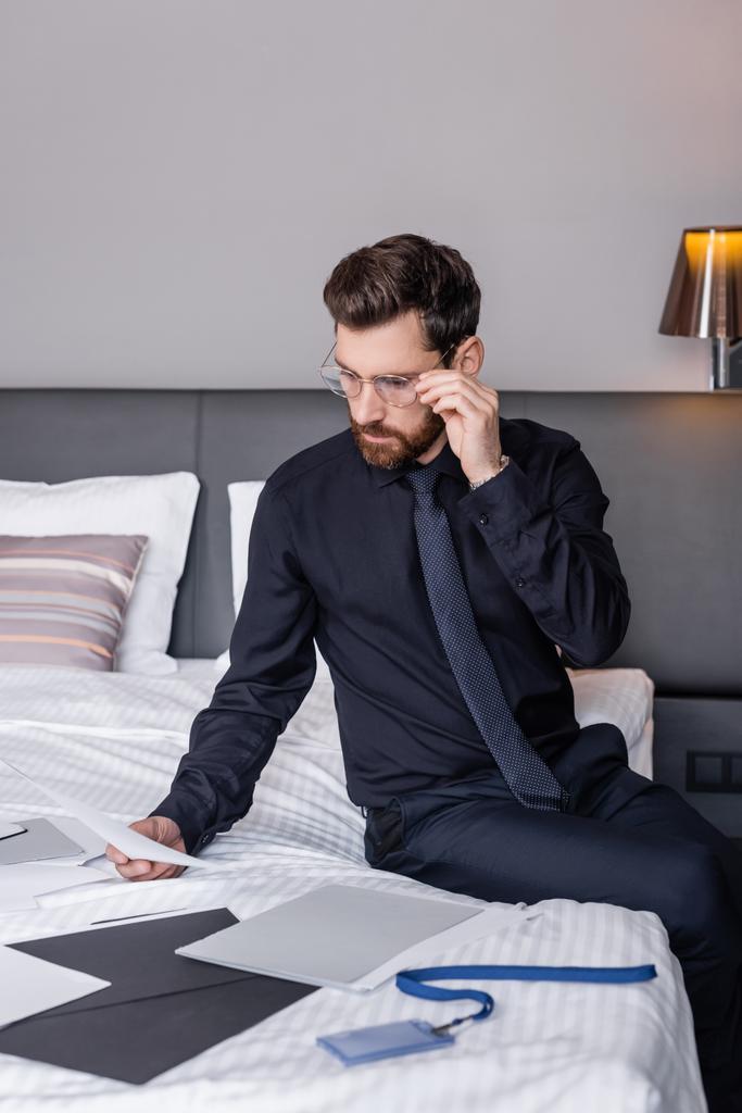 hombre barbudo de traje ajustando anteojos y mirando documentos en la cama del hotel  - Foto, Imagen