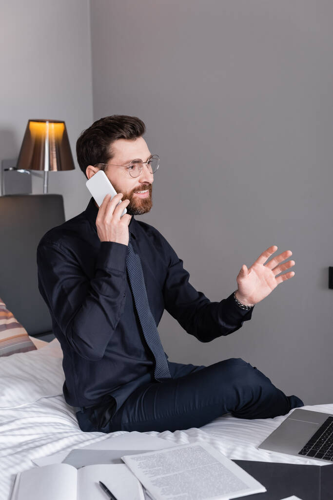 Χαρούμενος επιχειρηματίας με κοστούμι και γυαλιά που μιλάει στο smartphone του ξενοδοχείου  - Φωτογραφία, εικόνα